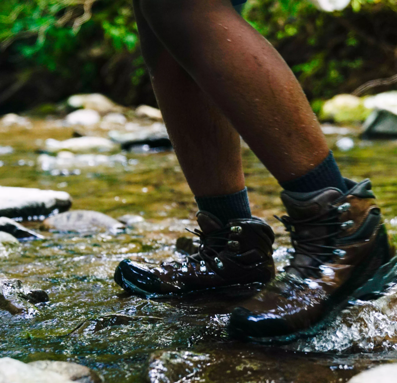 The image shows a person wading through a shallow stream or river. The focus is on their lower legs and feet, which are clad in hiking boots and dark socks. The water is clear and reflects the surrounding environment, with rocks visible both in and out of the water. The person's skin is dark, and their legs appear wet. The overall impression is one of outdoor activity and immersion in nature.